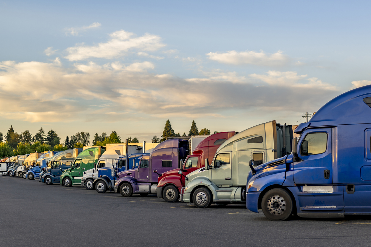 Commercial haulers big rig semi trucks tractors and semi trailers standing in row on truck stop parking lot with sunset clouds sky take a break according to log book waiting for the next loads