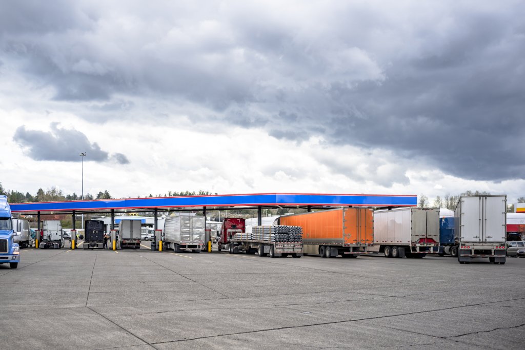 Industrial carriers different big rig semi trucks tractors with loaded semi trailers standing in row on the truck stop gas station filling up tanks to be able to continue the freights