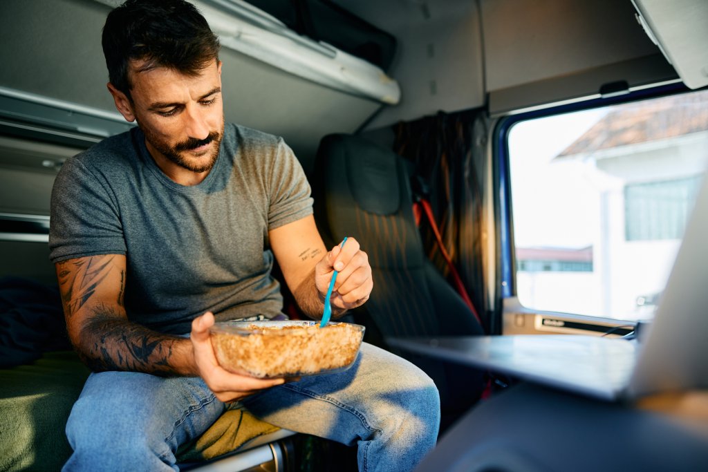 Mid adult driver eating in truck cabin while resting on a parking lot.